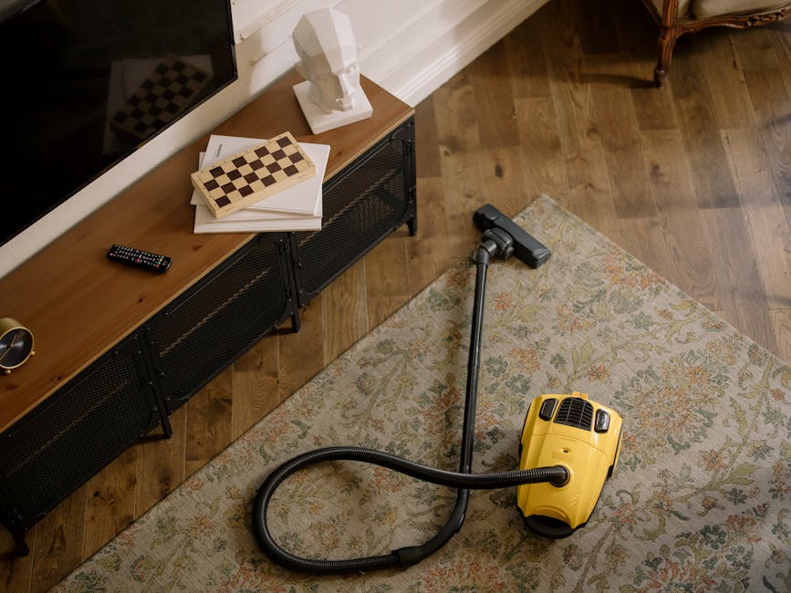 A person kneeling on a patterned carpet in a living room, using a yellow and black vacuum cleaner to perform surface cleaning. The vacuum's dust container and hose are visible, and the individual is p
