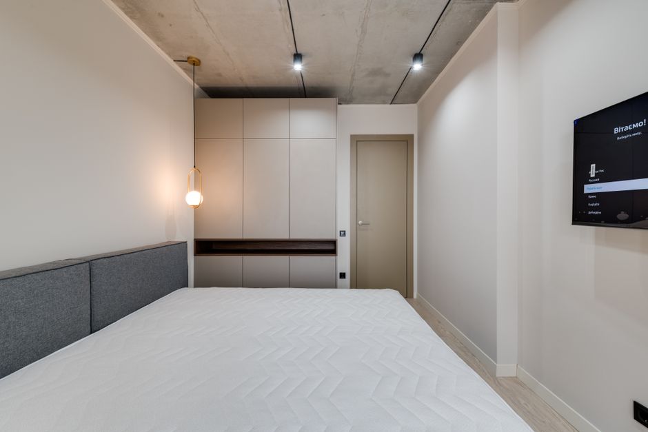 A minimally decorated bedroom featuring a white upholstered bed with a clean, made-up mattress, positioned against a white wall. To the right of the bed, a modern beige wardrobe with sleek, vertical h